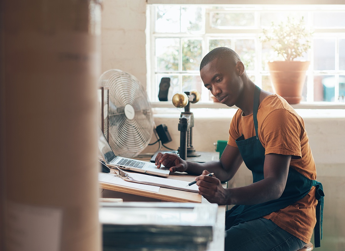 Business Insurance - Portrait of a Young Small Business Owner Wearing an Apron Working on a Laptop While Sitting Behind a Desk in his Bright Office