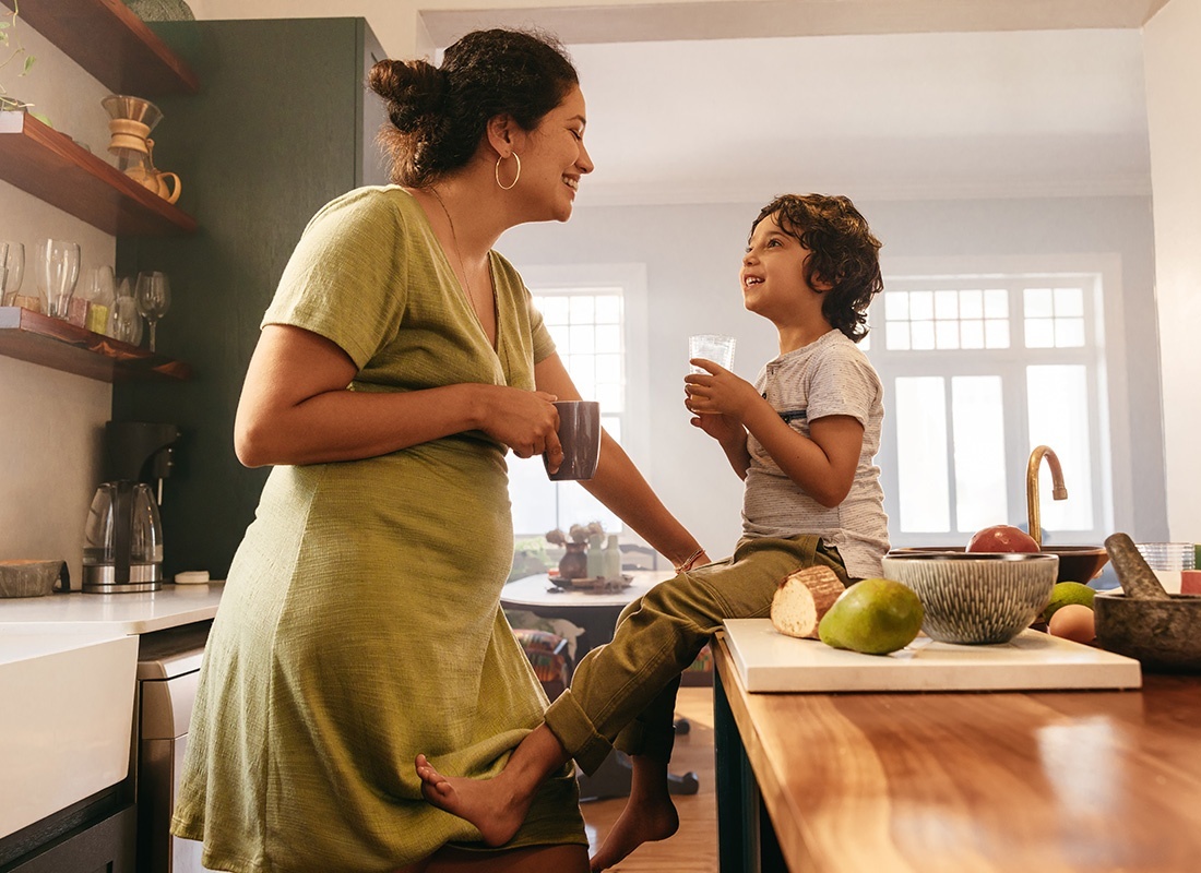 Individual Life Insurance - Portrait of a Cheerful Young Mother Holding a Cup of Coffee While Standing in the Kitchen Looking at her Younger Son Sitting on the Kitchen Island
