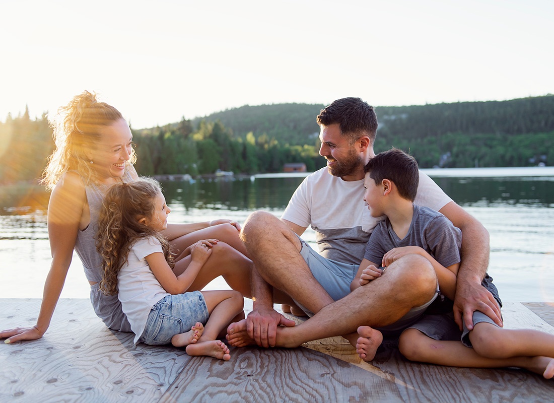 Personal Insurance - Portrait of a Cheerful Family with Two Young Kids Having Fun Sitting on a Wooden Dock by the Lake at Sunset While on Vacation